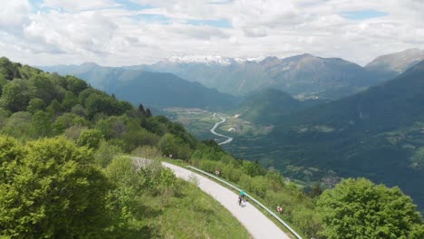 cyclists-riding-mountain-road-above-picturesque-valley,-paraglider-flies-by,-aerial