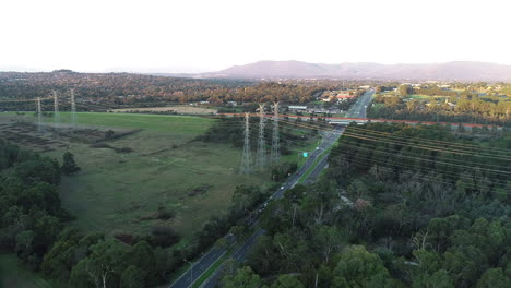 Suspended-power-lines-linked-above-green-belt-highway-and-road-infrastructure-with-beautiful-mountain-view-in-the-distance