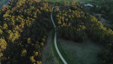 Smooth-drone-view-of-park-pathway-beside-busy-highway-and-sporting-arena