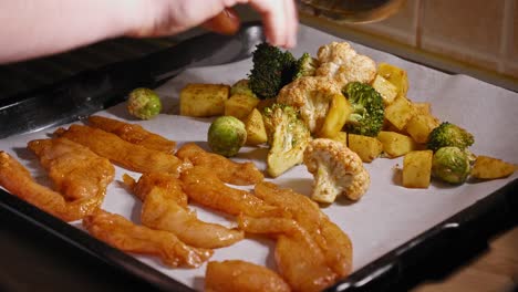 A-close-up-shot-of-a-baking-tray-filled-with-marinated-chicken-strips,-as-an-assortment-of-fresh-vegetables-are-added-onto-the-tray-before-a-chef-carefully-spreads-the-pieces-to-ensure-an-even-roast