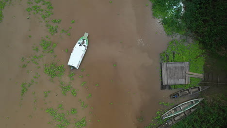 Cinematic-rising-drone-shot-of-boat-leaving-a-dock-in-the-amazon-river,-with-lush-amazon-rainforest-surrounding-the-water-in-Peru