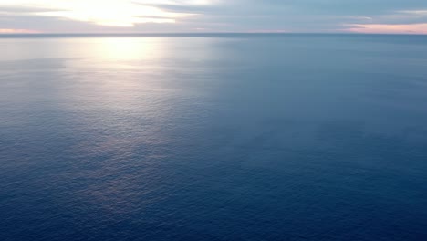 An-endless-blue-ocean-stretching-out-to-the-horizon-at-dusk---aerial-pull-back-flyover