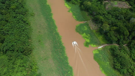 Cinematic-drone-shot-with-a-boat-coming-down-the-Amazon-river-and-the-rainforest-surrounding-the-river-in-Peru,-slowly-tilting-downward-towards-the-boat