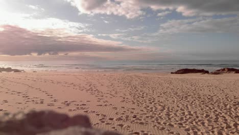 Goldener-Sandstrand-Mit-Fußspuren-Bedeckt,-Sonnenuntergang-Beleuchtet-Wolken-über-Dem-Meer