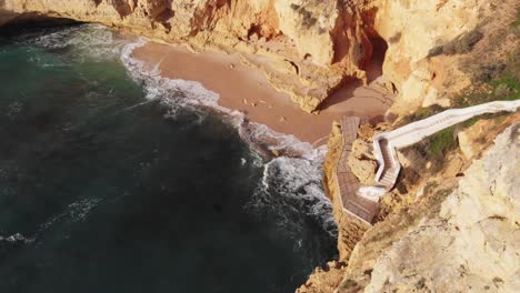 Aerial-drone-shot-of-high-waves-crashing-against-the-beach-with-steep-high-cliffs-on-all-sides-and-white-stairways-leading-upto-the-beach-at-sunset