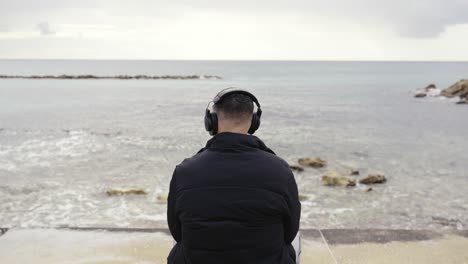Ein-Mann-Sitzt-Auf-Einer-Bank-Am-Meer-Und-Hört-Mit-Seinen-Kopfhörern-Musik