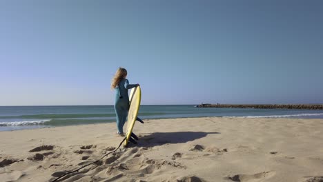 Young-girl-wearing-wetsuit-and-picking-up-surfboard-on-beach,-getting-ready-for-surfing