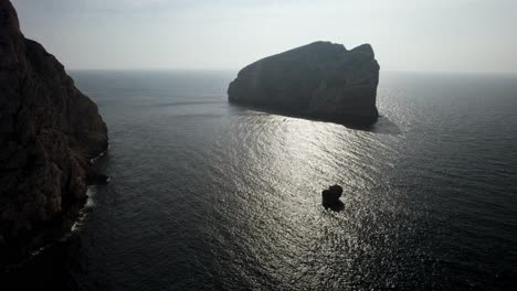 Aerial-establishing-shot-of-the-silhouette-of-the-huge-rock-formation-against-the-sun-reflecting-on-the-surface-of-the-sea