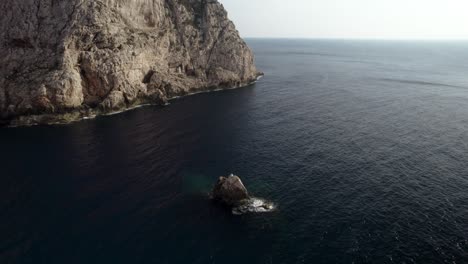 Aerial-shot-of-the-small-rock-and-huge-cliffs-and-a-dark-calm-Mediterranean-sea-surface