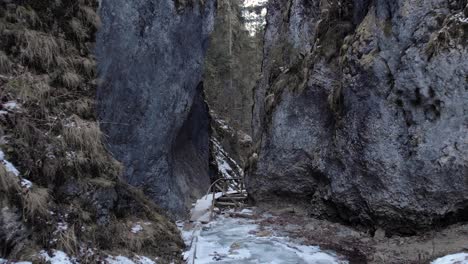Backward-Through-Narrow-Gap-Between-Rugged-Cliffs-With-Wooden-Bridge-In-Winter-Forest
