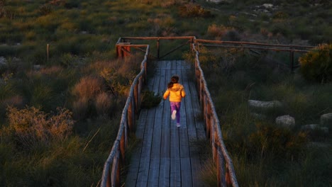 Adorable-young-girl-running-along-a-boardwalk-to-go-to-the-beach-in-winter-at-sunset