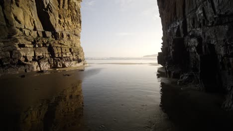 A-huge-sea-cave-with-tide-water-coming-through-during-sunset-by-the-ocean-with-light-reflected-on-the-water-above-the-sand