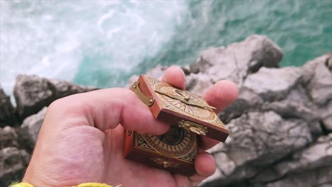 Close-up-of-human-hand-opening-in-slow-motion-a-wooden-engraved-compass-in-a-rocky-coastal-landscape