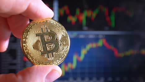 Male-finger-holding-golden-Bitcoin-in-light-and-stock-index-curves-in-background,close-up