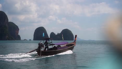 Traditional-Thai-boat-sails-at-high-speed-away-from-a-tourist-attraction-over-a-calm-sea-on-a-partly-cloudy-day