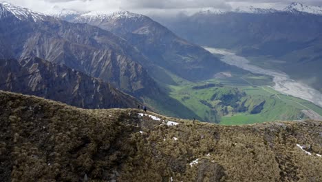 Hikers-on-a-beautiful-ridge-overlooking-huge-mountains-and-green-valley-with-a-river-running-through-in-New-Zealand-on-a-bright-sunny-day