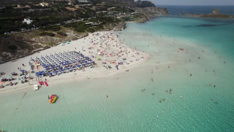 Aerial-establishing-orbit-shot-of-one-of-the-most-beautiful-beaches-on-the-Emerald-Coast-in-Sardinia-with-people-sunbathing-and-swimming-in-the-crystal-clear-waters