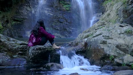 Mädchen-Sitzt-Auf-Felsen-Mit-Fließendem-Wasser-Des-Wasserfalls-Vom-Berg-Im-Wald-Aus-Einem-Video-Mit-Flachem-Winkel,-Das-Am-Thangsingh-Wasserfall-Shillong-Meghalaya-Indien-Aufgenommen-Wurde