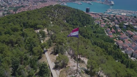 Flying-around-the-flag-of-Croatia,-keeping-it-as-the-main-subject-and-the-port-and-city-of-Split-in-the-background