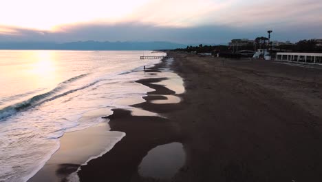 Bewölkter-Sonnenuntergang-über-Dem-Strand,-Kleine-Wellen-Und-Mädchen,-Die-Vor-Dem-Wasser-Stehen,-Drohnenaufnahme