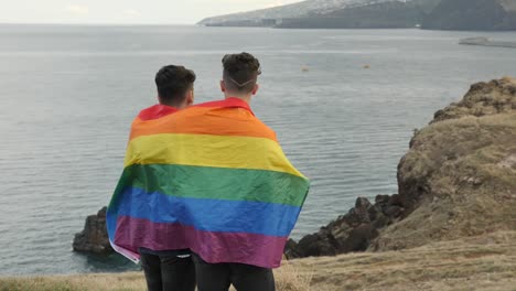 Gay-couple-wrapped-in-a-pride-flag-looking-into-a-greater-future,-homosexual-standing-in-the-side-of-a-cliff