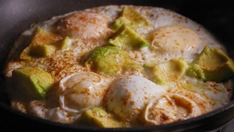 A-close-up-shot-of-a-frying-pan-cooking-eggs-with-avocado-covered-in-light-seasoning,-the-lid-from-the-pan-removed-and-hot-steam-escapes-as-a-chef-prepares-the-ingredients-to-create-an-open-sandwich