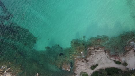 Aerial-top-down-shot-of-a-coastline,-emerald-sea,-and-a-sandy-beach-completely-empty-due-to-the-coronavirus-pandemic-lockdown-in-Porto-Cervo,-Sardinia,-Italy
