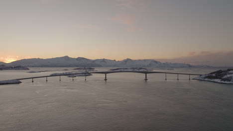 Beautiful-aerial-shot-of-a-bridge-over-water-in-snowy-mountains-at-sunset