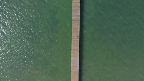 Top-Down-Aerial-View-as-Tourists-Walk-on-Wooden-Fishing-Pier-with-Emerald-Green-Waters,-El-Muellecito,-Monte-Cristi,-Dominican-Republic
