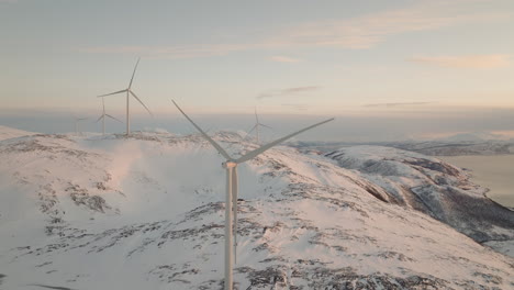 Aerial-snowy-mountain-view-of-wind-turbine-blades-turning,-arctic-sunset