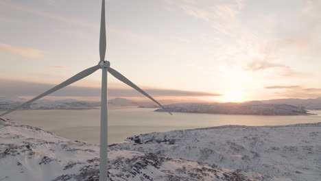 Wind-turbine-slowly-turns-on-white-Arctic-mountain,-vivid-sunset-over-fjord
