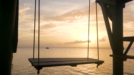 An-empty-wooden-swing-is-swinging-on-a-dock-overlooking-the-ocean-with-a-boat-in-the-orange-rays-of-the-sunset