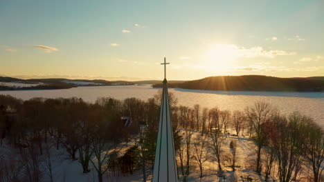 Scenic-aerial-view-of-a-church-steeple-against-a-winter-sunset-across-a-frozen-lake