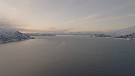 Cargo-ship-in-fjord-at-sunset,-white-winter-landscape-in-Norway