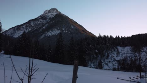 Vuelo-Panorámico-Sobre-La-Nieve-Hacia-Una-Enorme-E-Impresionante-Montaña-Con-Bosques-Y-árboles-En-Invierno-Con-Nieve-Y-Hielo-En-Las-Idílicas-Montañas-Bávaras-De-Los-Alpes-Austríacos-Con-Picos-Rojos-Al-Atardecer