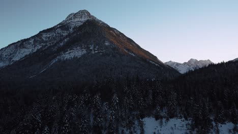 Flying-along-a-huge-and-impressive-mountain-with-forest-and-trees-in-winter-with-snow-and-ice-in-the-idyllic-and-scenic-Bavarian-Austrian-alps-mountains-with-red-sunset-peaks