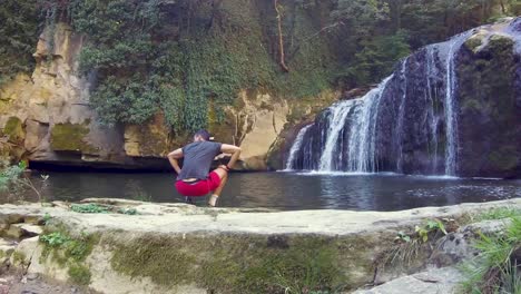 Boy-rise-from-squat-position-with-majestic-waterfall-view-in-front-of-him,-static-back-shot