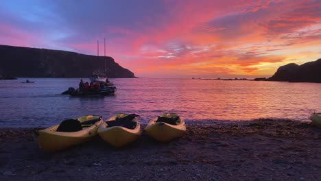 Kajaks-Ruhen-Sich-Bei-Sonnenuntergang-Am-Strand-Von-Catalina-Aus,-Während-Menschen-Auf-Anderen-Schiffen-Segeln