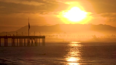 Ein-Surfer-Paddelt-Bei-Morgendämmerung-Oder-Sonnenuntergang-Auf-Den-Ozean-Mit-Dem-Ventura,-Dem-California-Pier-In-Der-Ferne-Und-Den-Heranrollenden-Wellen