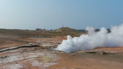 Aerial-Past-Steaming-Hot-Fumeroles-In-A-Geothermal-Aera-Reveals-The-Pretty-Reykjanes-Lighthouse-In-The-Distance
