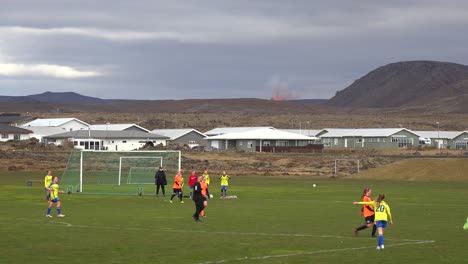 El-Volcán-Fagradalsfjall-Se-Ve-En-Erupción-En-La-Distancia-Detrás-De-Un-Partido-De-Fútbol-Femenino-En-La-Ciudad-De-Grindavik,-Islandia