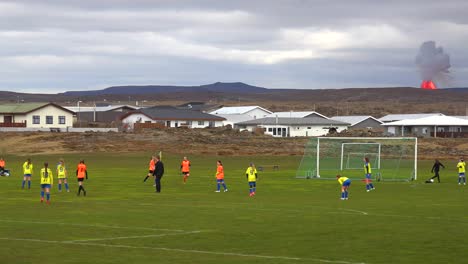 The-Fagradalsfjall-Volcano-Is-Seen-Erupting-In-The-Distance-Behind-A-Girls-Soccer-Match-The-Town-Of-Grindavik,-Iceland