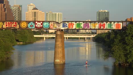A-Paddleboarder-Rows-Down-The-Colorado-River-In-Front-Of-The-Skyline-Of-Downtown-Austin-Texas
