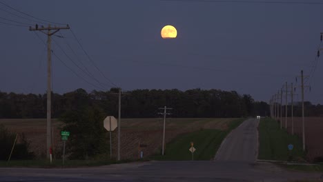 A-Distant-Car-Drives-Down-A-Lonely-Country-Road-At-Night-With-A-Full-Moon-Rising