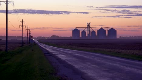 Grain-Silos-On-A-Large-Commercial-Farm-Along-A-Country-Road-At-Dusk-In-The-Midwest