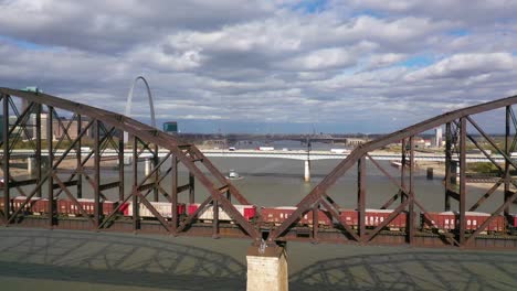 Aerial-Of-A-Freight-Train-Crossing-The-Mississippi-River-With-The-St-Louis-Gateway-Arch-And-City-Skyline-Background