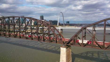 Aerial-Of-A-Freight-Train-Crossing-The-Mississippi-River-With-The-St-Louis-Gateway-Arch-And-City-Skyline-Background