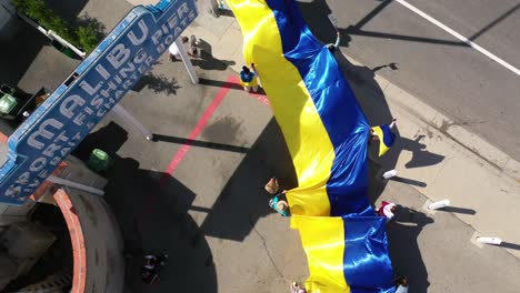 Ukrainische-Demonstranten-Aus-Der-Luft-Von-Oben-Nach-Unten-Mit-Großer-Ukrainischer-Flagge-Protestieren-Gegen-Die-Russische-Invasion-Ihres-Landes,-Auf-Dem-Malibu-Pier,-Kalifornien-Mit-Malibu-Schild