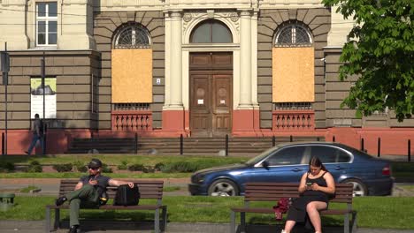 Ukrainians-Sit-On-A-Park-Bench-Enjoying-The-Sunshine-In-Central-Lviv,-Ukraine-With-Boarded-Up-Buildings-In-Background