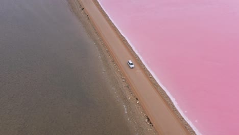Excellent-Aerial-Shot-Of-Of-A-Car-Driving-Down-A-Road-Dividing-Lake-Macdonnell-On-Eyre-Peninsula,-South-Australia,-With-Brown-Water-On-One-Side-And-Pink-On-The-Other